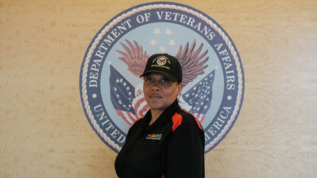 A Black female VA employee, wearing a “Department of Veterans Affairs” baseball cap, stands in front of a VA seal mural on the wall of a VA facility.