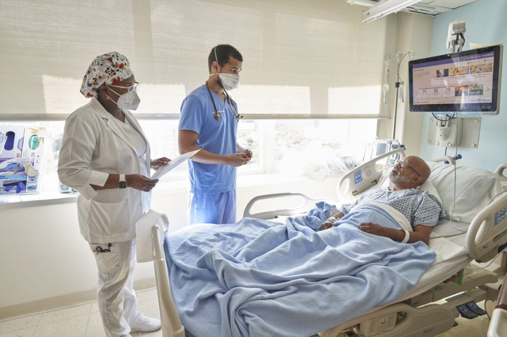 A Veteran patient lies in a hospital bed while two health care professionals provide attentive care. At VA, it’s all about Veterans—ensuring they receive quality medical support.