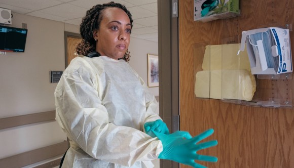 A female physician in protective gear dons gloves as she enters a patient’s room at a VA facility.